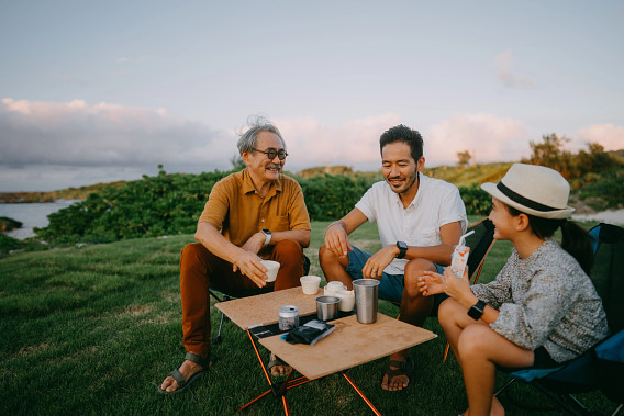 This image shows three people sitting outdoors on a grassy area, enjoying a casual gathering. They are seated around a small portable table with drinks and cups, smiling and engaging in conversation. The background features lush greenery and an open sky, suggesting a relaxed and pleasant environment, possibly during a picnic or camping trip.