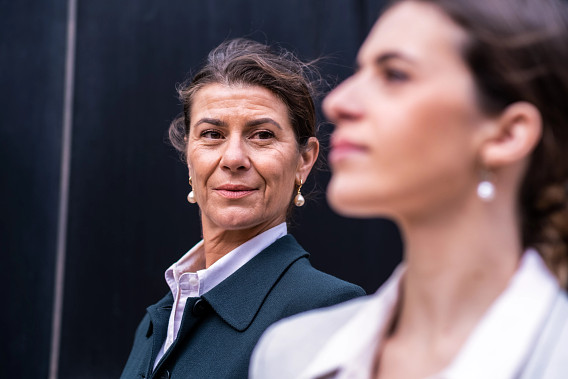 This image shows two women in professional attire, with one in focus and the other blurred in the foreground. The woman in focus, wearing pearl earrings and a confident expression, appears to be looking toward the other. The setting suggests a formal or business-like environment, highlighting themes of mentorship and leadership.