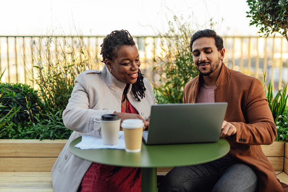 Two colleagues sitting outdoors at a green table, working together on a laptop. Both are smiling and engaged in conversation, with coffee cups and documents on the table. The setting includes plants and a railing, suggesting a relaxed, professional environment.