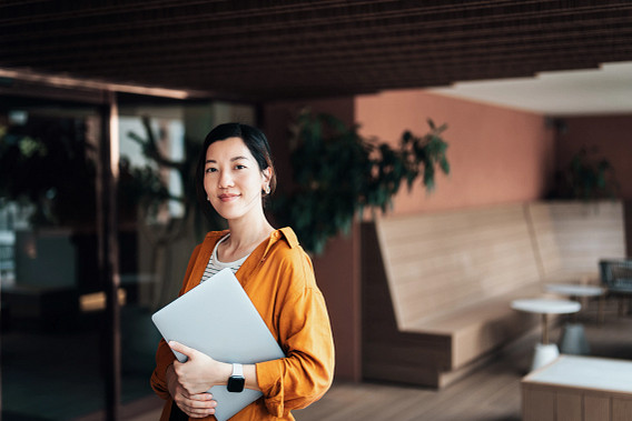 A lady in mustard yellow, looking into the camera with a laptop in her arms at an office open area.