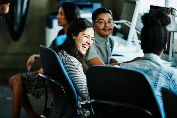 The image shows a group of diverse people, including a smiling woman, sitting in what appears to be an office or work environment. The people are dressed in casual or business attire, and the image conveys a sense of collaboration and engagement.